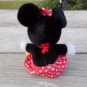 May include: A plush Minnie Mouse doll, viewed from the back, with black fur, large ears, and a red bow. The doll wears a red skirt with white polka dots, white gloves, and a red bow at the waist.