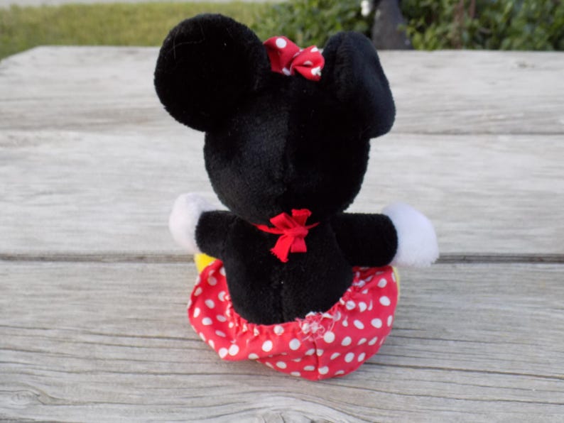 May include: A plush Minnie Mouse doll, viewed from the back, with black fur, large ears, and a red bow. The doll wears a red skirt with white polka dots, white gloves, and a red bow at the waist.