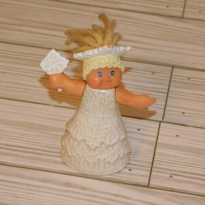 May include: A vintage doll with blonde yarn hair, blue eyes, and a white dress. The doll is holding a white snowflake-shaped object. The doll's skin is a peach color. The doll is standing on a light brown wooden surface.