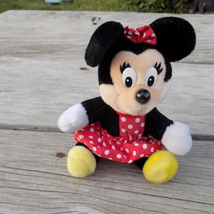 May include: A plush Minnie Mouse doll, featuring black ears, a red and white polka dot dress, and a matching bow. The doll has white gloves and yellow shoes, and is sitting on a wooden surface.
