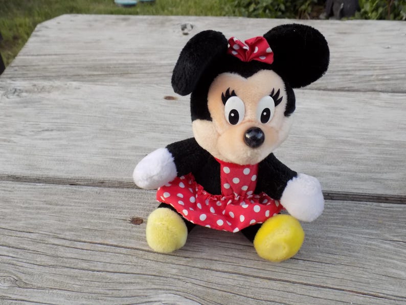 May include: A plush Minnie Mouse doll, featuring black ears, a red and white polka dot dress, and a matching bow. The doll has white gloves and yellow shoes, and is sitting on a wooden surface.