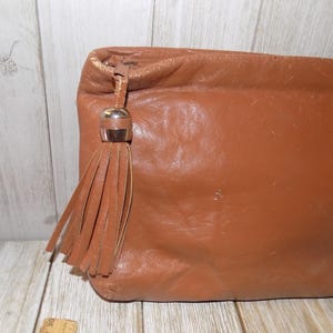 May include: A brown leather clutch bag with a tassel and a gold-toned metal accent. The bag has a soft, textured surface.