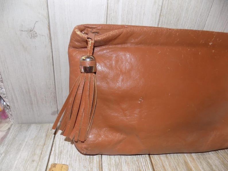 May include: A brown leather clutch bag with a tassel and a gold-toned metal accent. The bag has a soft, textured surface.