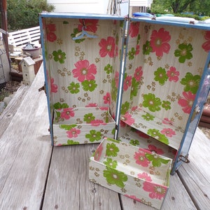 May include: A vintage blue metal dollhouse wardrobe with a floral print interior. The wardrobe has a hanging rack, two shelves, and a pull-out drawer. The floral print features pink and green flowers on a cream background.
