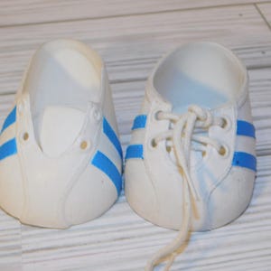 May include: Pair of white sneakers with blue stripes. The shoes have a classic design with laces and a rounded toe. The blue stripes are on the sides of the shoes. The shoes are made of a smooth material.