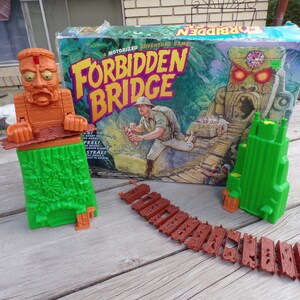 May include: A vintage board game called "Forbidden Bridge" with a green and brown jungle theme. The box art features a cartoon explorer and a stone idol with glowing red eyes. The game includes a green tower, a brown bridge, and a brown idol with a green base.