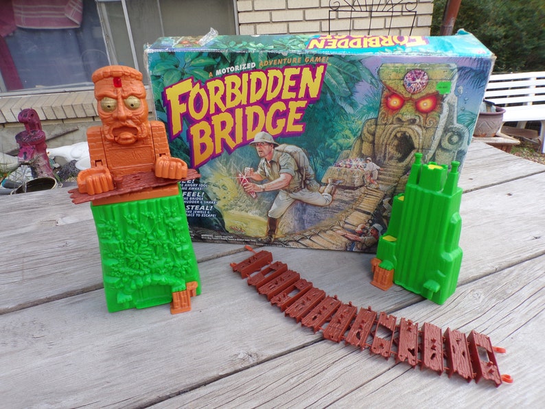May include: A vintage board game called "Forbidden Bridge" with a green and brown jungle theme. The box art features a cartoon explorer and a stone idol with glowing red eyes. The game includes a green tower, a brown bridge, and a brown idol with a green base.
