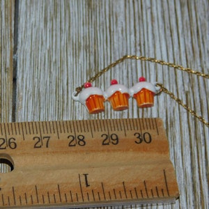 May include: A gold chain necklace with three cupcake charms. The cupcakes are white with orange frosting and pink sprinkles.
