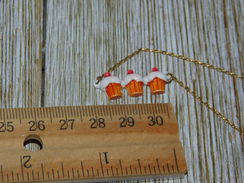 May include: A gold chain necklace with three cupcake charms. The cupcakes are white with orange frosting and pink sprinkles.