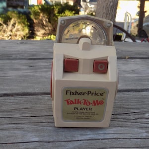 May include: A vintage Fisher-Price Talk-To-Me Player toy in cream and red. The toy has a clear plastic dial and two red buttons. The label reads "Fisher-Price Talk-To-Me PLAYER". The toy is sitting on a weathered wooden surface.