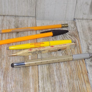 May include: A collection of writing instruments including pencils, pens, and a stylus. The pens are yellow and orange, with black ink. One pen has the text "AUTO SERVICE CENTERS KEARNS 4015 West 5400 So. KILN DRIED".