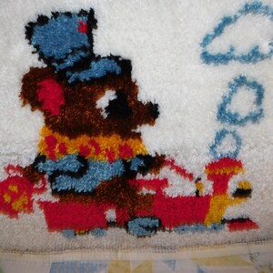 May include: A brown bear wearing a blue hat and a yellow and red scarf is driving a red and yellow toy car. The bear is puffing out smoke from the car's chimney. The image is a rug made of yarn.