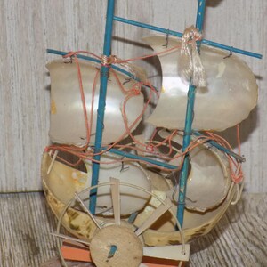 May include: A miniature sailboat model crafted from seashells, featuring blue masts and orange rigging. The sails are made from translucent shells, and a wooden wheel is visible at the base. The model is a decorative nautical item.