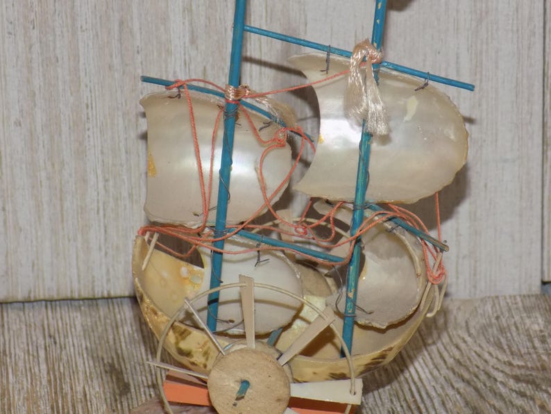 May include: A miniature sailboat model crafted from seashells, featuring blue masts and orange rigging. The sails are made from translucent shells, and a wooden wheel is visible at the base. The model is a decorative nautical item.