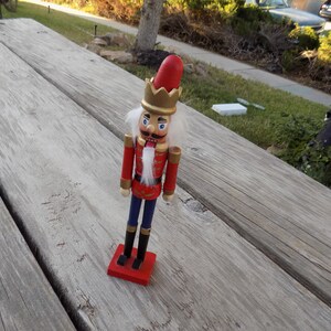 May include: A wooden nutcracker figurine with a red and gold uniform, a white beard, and blue pants. The nutcracker is standing on a red base.