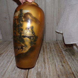 May include: A gold ceramic vase with a Japanese-inspired design featuring a mountain landscape, a small house, and a horse. The vase has a glossy finish and is decorated with black and white details.