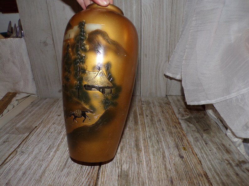 May include: A gold ceramic vase with a Japanese-inspired design featuring a mountain landscape, a small house, and a horse. The vase has a glossy finish and is decorated with black and white details.