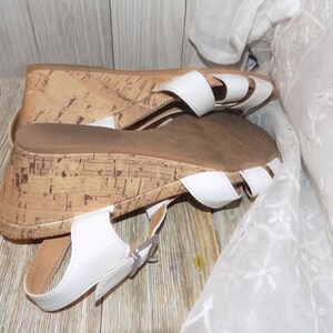 K&ouml;nnte beinhalten: Wei&szlig;e Keilsandalen mit Korksohlen und einem Schnallenverschluss. Die Sandalen haben einen Riemen &uuml;ber den Zehen und einen Riemen um den Kn&ouml;chel.