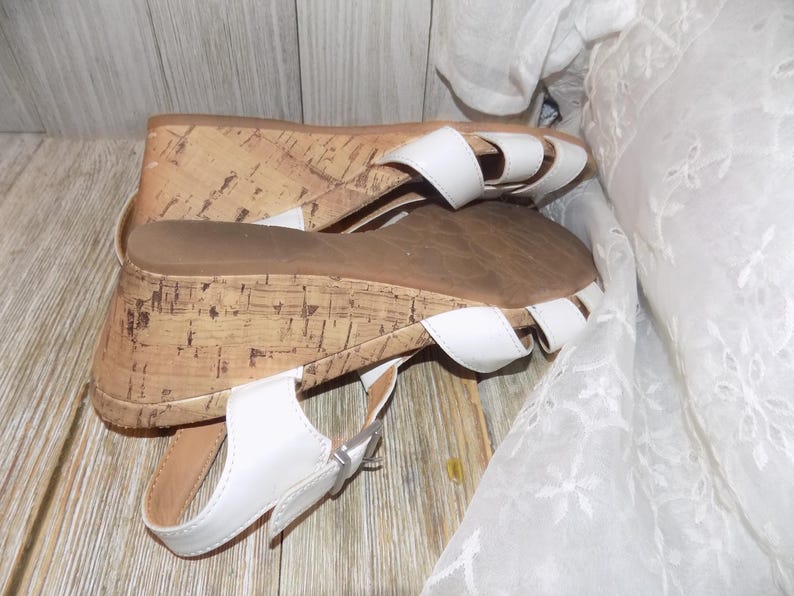 K&ouml;nnte beinhalten: Wei&szlig;e Keilsandalen mit Korksohlen und einem Schnallenverschluss. Die Sandalen haben einen Riemen &uuml;ber den Zehen und einen Riemen um den Kn&ouml;chel.