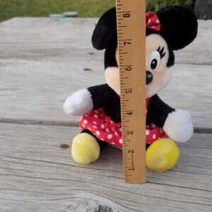 May include: A Minnie Mouse plush toy, approximately 8 inches tall, with black ears, a red and white polka dot dress, and yellow feet. The toy has white gloves and a red bow. The plush is sitting on a wooden surface.