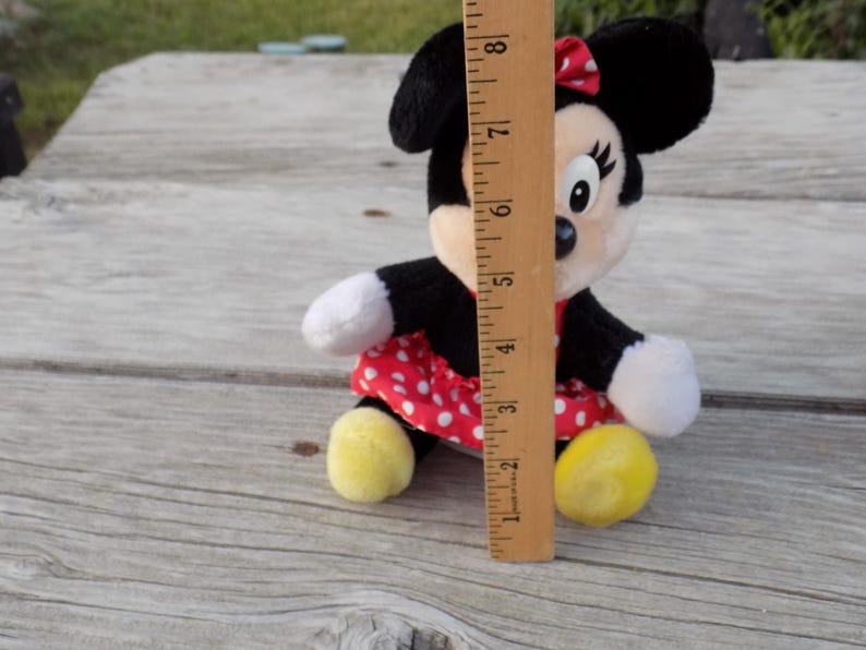 May include: A Minnie Mouse plush toy, approximately 8 inches tall, with black ears, a red and white polka dot dress, and yellow feet. The toy has white gloves and a red bow. The plush is sitting on a wooden surface.