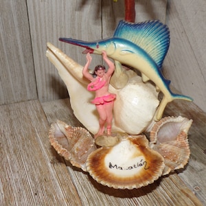 May include: A vintage souvenir from Tahiti featuring a plastic figurine of a hula dancer standing on a seashell with a blue and yellow marlin fish jumping over her. The base of the figurine is made of several seashells with the word "Ma-atiá" written on one of them.