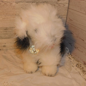 May include: A fluffy white and black stuffed animal dog with a gold-colored medallion around its neck. The plush toy has a shaggy appearance and is sitting on a white surface.