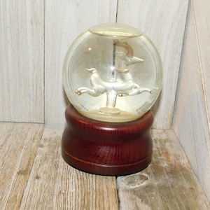 May include: A glass snow globe with a silver deer figurine inside. The globe sits on a dark wood base.
