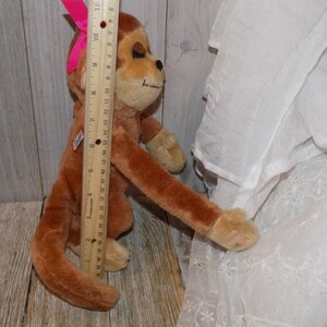 May include: A brown plush monkey toy with a pink bow on its head. The monkey is approximately 24 inches tall. It has tan paws and a tan face. A wooden ruler is placed next to the monkey.