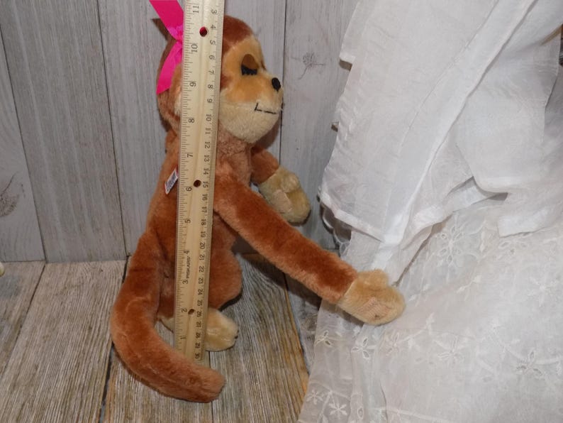 May include: A brown plush monkey toy with a pink bow on its head. The monkey is approximately 24 inches tall. It has tan paws and a tan face. A wooden ruler is placed next to the monkey.