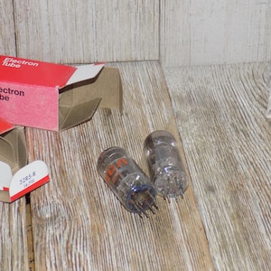 May include: Two clear glass RCA Electron Tubes with orange lettering and metal pins, next to a red and white box with the words "Electron Tube" printed on it. The tubes are labeled "32RS-R".