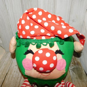 May include: A whimsical Christmas elf doll with a green body, a red and white polka-dot hat, and a matching nose. The elf has black eyelashes, pink cheeks, and a green trim with small green pom-poms. The elf's legs are red and white striped.