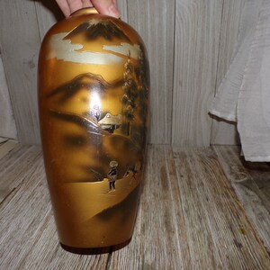 May include: A gold ceramic vase with a black and white painted scene of a mountain, trees, and figures. The vase has a glossy finish.