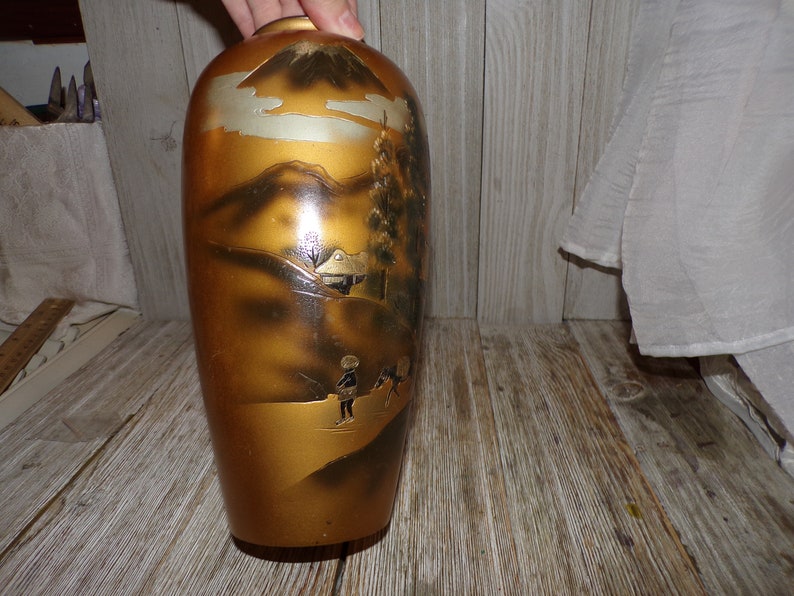 May include: A gold ceramic vase with a black and white painted scene of a mountain, trees, and figures. The vase has a glossy finish.