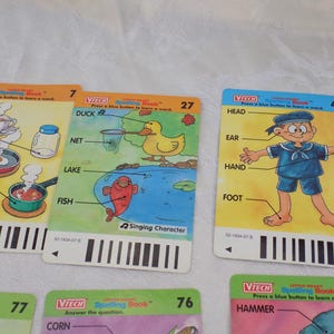May include: A set of rectangular VTech Little Smart Spelling Book cards featuring colorful illustrations and labels. Card 7 shows kitchen items, card 27 depicts a duck, net, lake, and fish, and card 45 shows a person with body parts labeled.