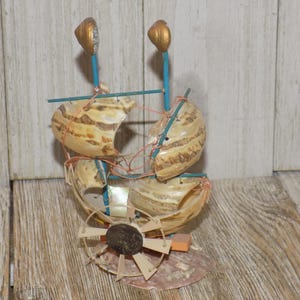 May include: A handcrafted nautical sculpture featuring a boat design made from seashells. The sculpture has a brown and beige color scheme, with blue accents and gold-colored finials. The boat is constructed with shells, wire, and wooden elements.