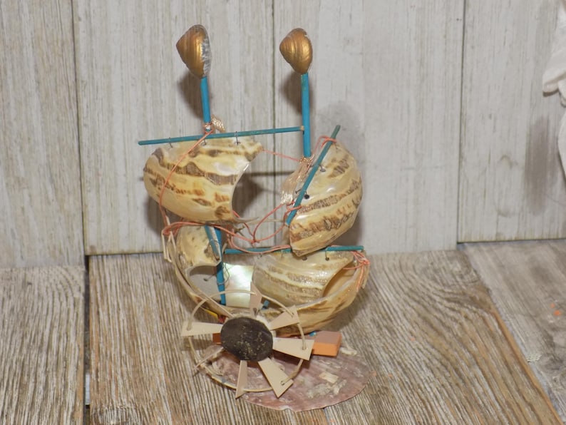 May include: A handcrafted nautical sculpture featuring a boat design made from seashells. The sculpture has a brown and beige color scheme, with blue accents and gold-colored finials. The boat is constructed with shells, wire, and wooden elements.