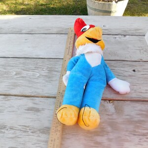 May include: A plush toy of Woody Woodpecker, a cartoon character. The toy is lying on a wooden surface and is wearing a blue jumpsuit with a white collar. The toy has a red head with a yellow beak and a white face with black eyes.