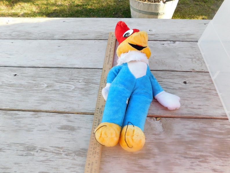 May include: A plush toy of Woody Woodpecker, a cartoon character. The toy is lying on a wooden surface and is wearing a blue jumpsuit with a white collar. The toy has a red head with a yellow beak and a white face with black eyes.
