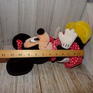 K&ouml;nnte beinhalten: Ein Pl&uuml;schtier von Minnie Mouse, das ein rotes Kleid mit wei&szlig;en Punkten und eine rote Schleife tr&auml;gt. Das Spielzeug liegt auf der Seite mit dem Kopf zur Seite gedreht. Das Spielzeug ist etwa 40 cm lang.
