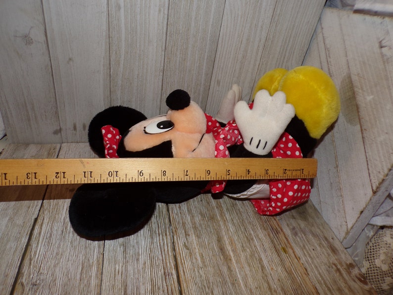 K&ouml;nnte beinhalten: Ein Pl&uuml;schtier von Minnie Mouse, das ein rotes Kleid mit wei&szlig;en Punkten und eine rote Schleife tr&auml;gt. Das Spielzeug liegt auf der Seite mit dem Kopf zur Seite gedreht. Das Spielzeug ist etwa 40 cm lang.