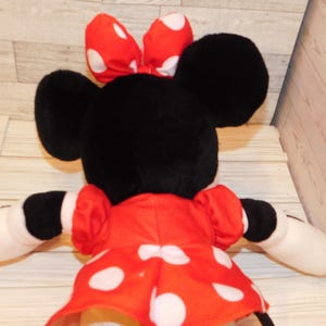 Op de afbeelding: Een pluche Minnie Mouse-pop, gezien van achteren, met zwarte oren en een rode strik met witte stippen. Ze draagt een rode jurk met witte stippen en heeft witte handschoenen.