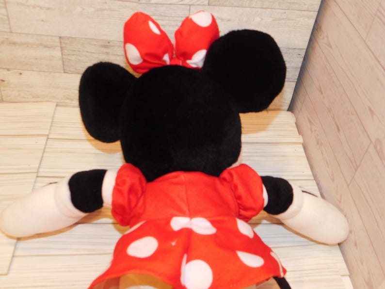 Op de afbeelding: Een pluche Minnie Mouse-pop, gezien van achteren, met zwarte oren en een rode strik met witte stippen. Ze draagt een rode jurk met witte stippen en heeft witte handschoenen.