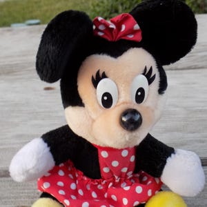 May include: A plush Minnie Mouse doll with black ears, a red and white polka dot bow, and a matching dress. The doll has white gloves and a yellow base. The doll is a popular character from Disney.