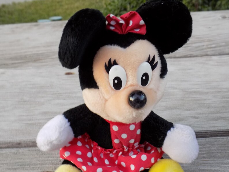May include: A plush Minnie Mouse doll with black ears, a red and white polka dot bow, and a matching dress. The doll has white gloves and a yellow base. The doll is a popular character from Disney.