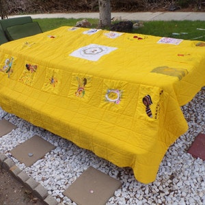 May include: A bright yellow quilt covers a rectangular table. The quilt features embroidered squares with various designs, including figures and floral patterns. The quilt is placed outdoors on a stone patio.