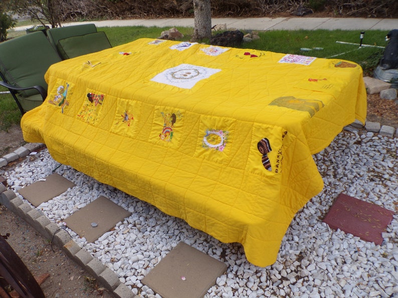 May include: A bright yellow quilt covers a rectangular table. The quilt features embroidered squares with various designs, including figures and floral patterns. The quilt is placed outdoors on a stone patio.