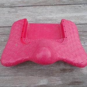 May include: Pink plastic children's play seat with a textured surface and a rounded center. The seat has two holes on each side.