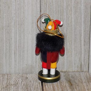May include: A wooden nutcracker ornament with a black fur collar, red and yellow clothing, and a multi-colored jester hat with a gold crown.