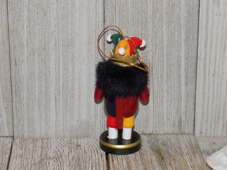 May include: A wooden nutcracker ornament with a black fur collar, red and yellow clothing, and a multi-colored jester hat with a gold crown.